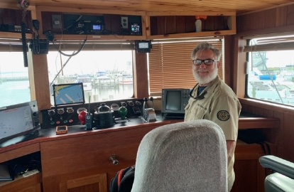 Captain Darien Hill standing at the helm console in a vessel's wheelhouse, with radar and instrument panels beside him and a marina visible through the windows.